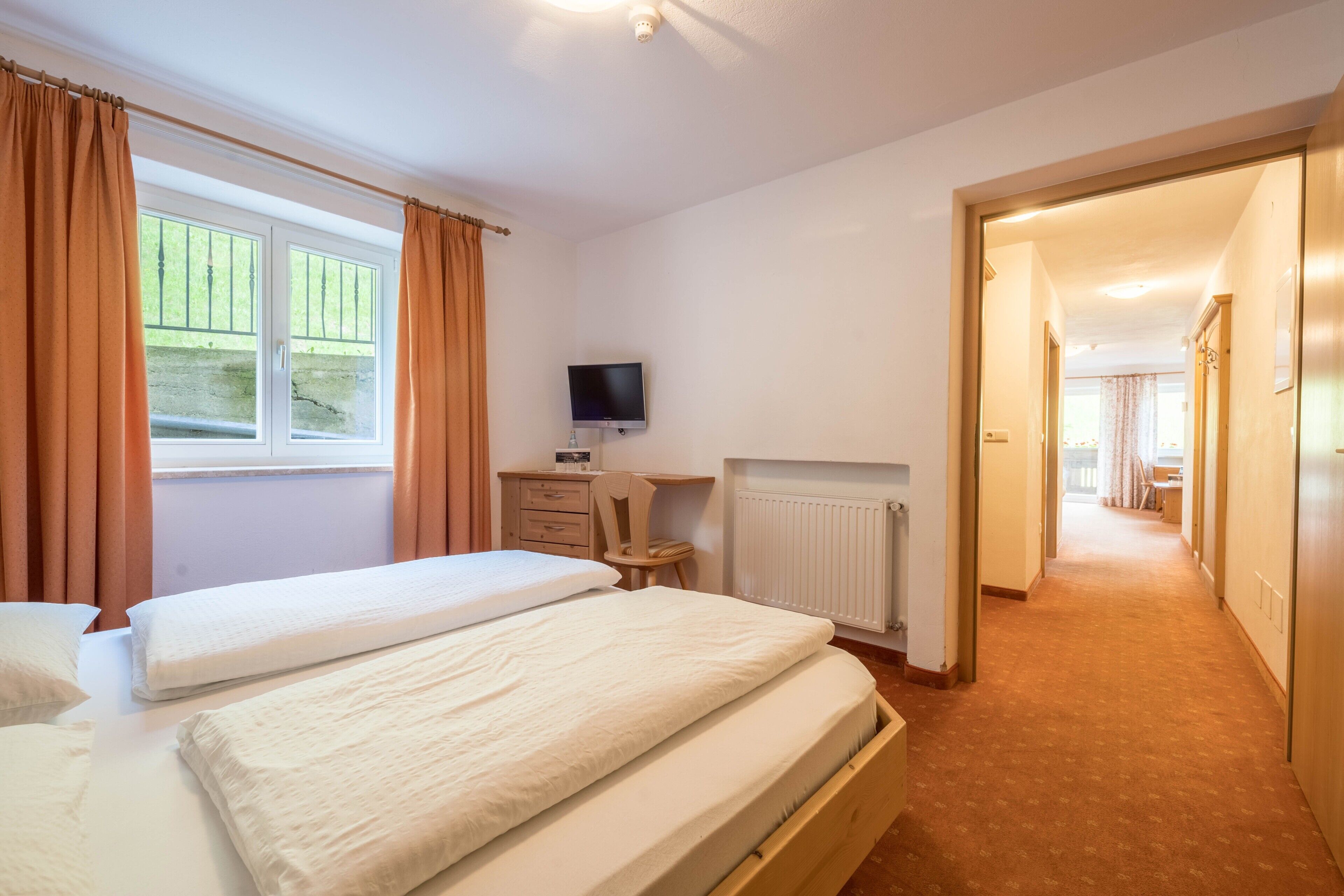 Slaapkamer met twee bedden, een bureau met computer, een radiator en uitzicht op een tuin door een raam.