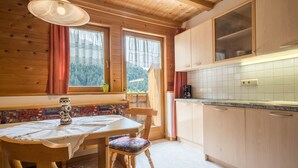 Interior de una cabaña de madera con una mesa de comedor, sillas, una ventana con cortinas, una zona de cocina con armarios y vistas al bosque.