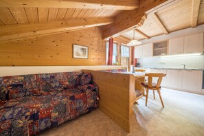 A cozy cabin interior with a wooden ceiling, a colorful sofa, a dining area, and a kitchen with white cabinets.