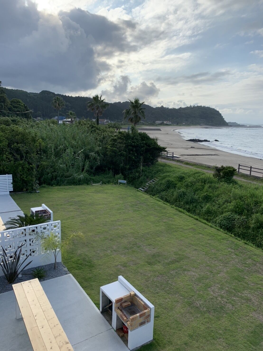 Uma área gramada com um banco de madeira e uma pequena construção branca, com vista para uma praia e colinas arborizadas.
