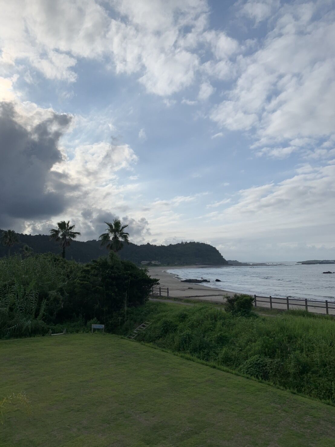 A coastal landscape with a grassy foreground, a beach, and a hilly backdrop under a partly cloudy sky.