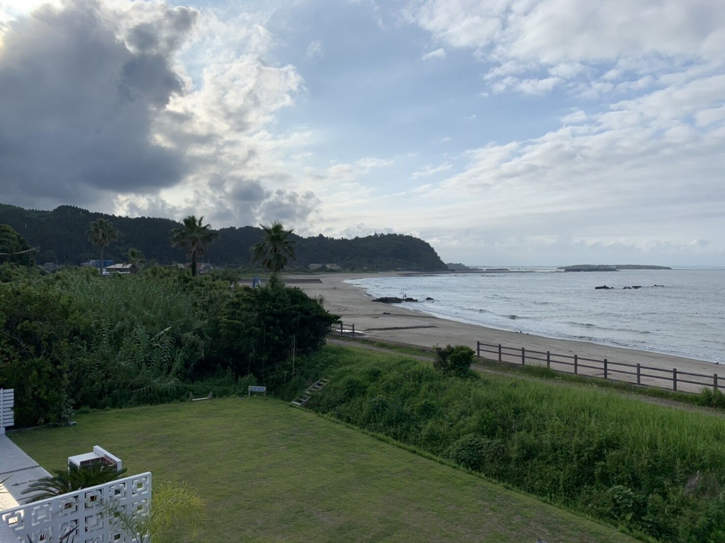 A coastal landscape with a beach, palm trees, and a grassy area with a fence.