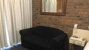 Une chambre d’hôtel avec un canapé noir, une petite table de chevet avec un téléphone et une horloge, un mur en briques et une fenêtre avec des rideaux blancs.