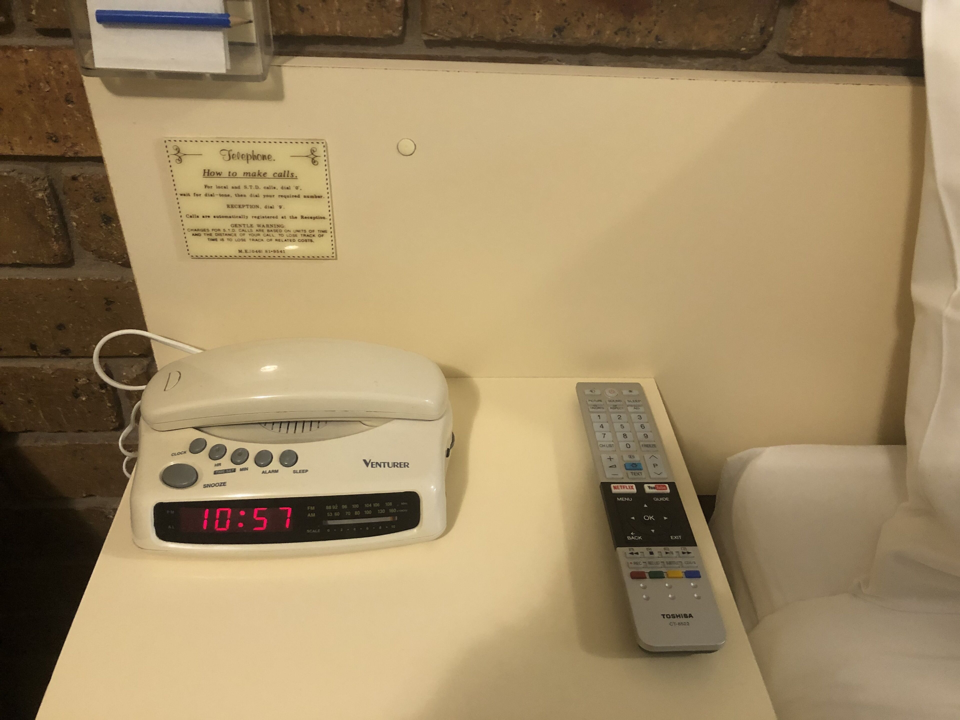 A white bedside table with a white clock displaying 10:57, a white Venturer phone, and a white remote control.