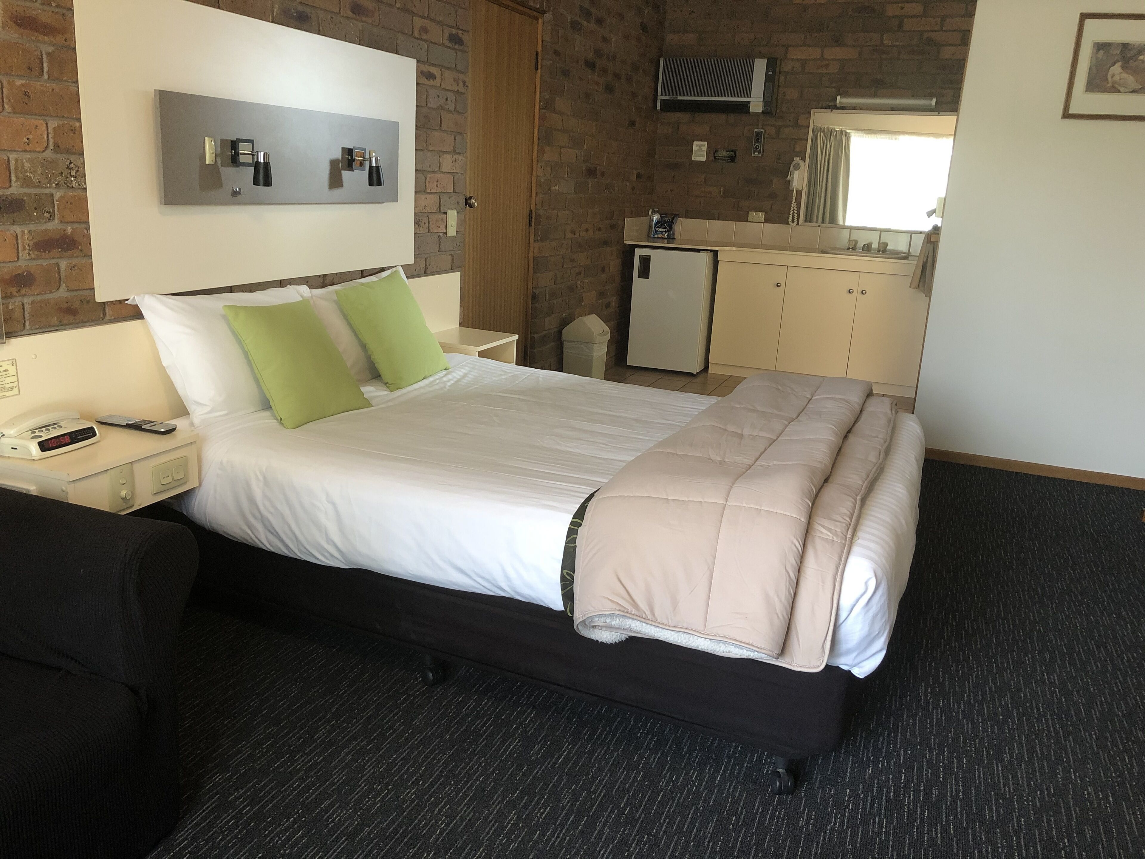 A hotel room with a bed, a nightstand with a phone, a small refrigerator, and a kitchenette area.