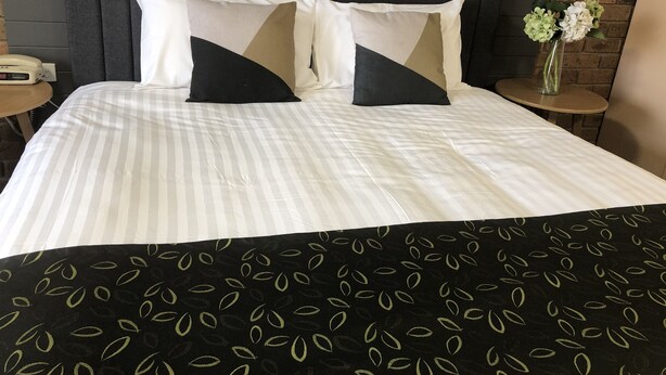 Un lit bien fait, avec une literie à rayures et deux oreillers, une petite table de chevet avec une lampe, et un vase rempli de fleurs posé sur un mur en briques.