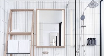 Un baño moderno con espejo de marco de madera, un perchero con toallas blancas, una ducha con cabezal de ducha manual y un dispensador de jabón de pared.