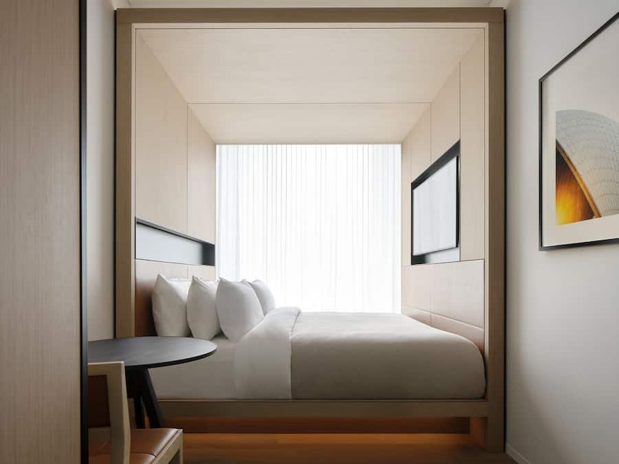 A modern hotel room with a bed, bedside table, and a window with blinds.