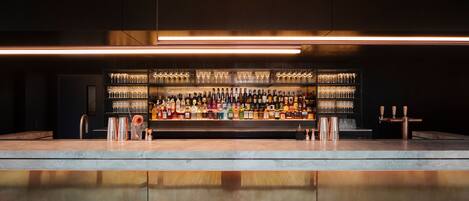 Una zona de bar con una barra alargada, estantes llenos de botellas y vasos, y un techo moderno de madera.