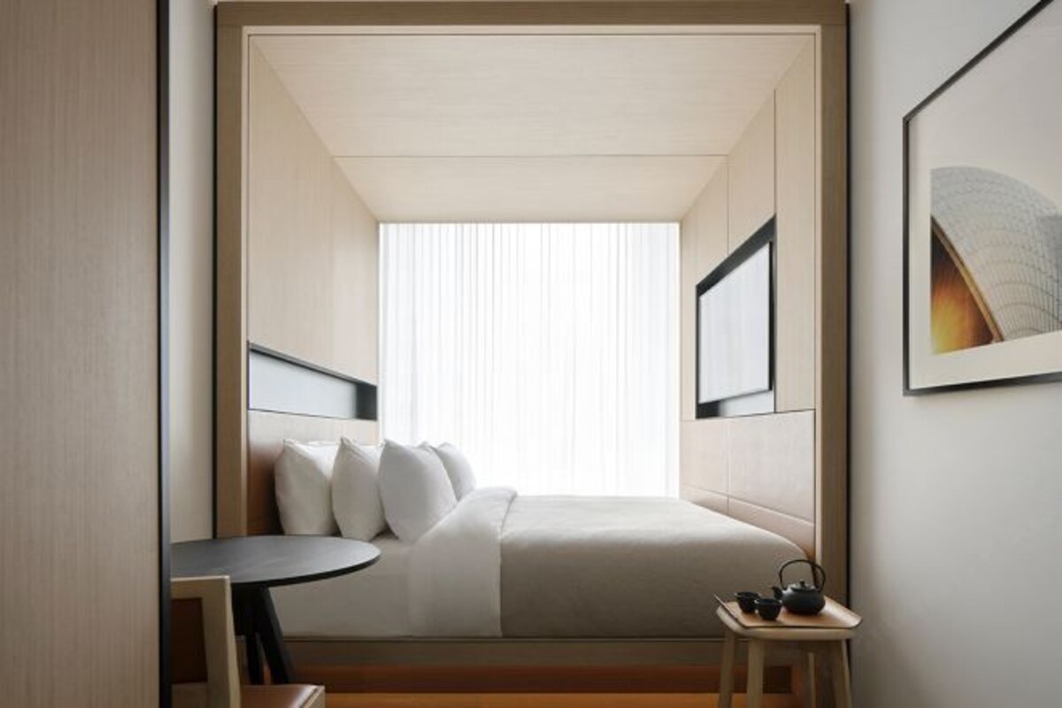 A hotel room with a bed, a small table, a chair, a TV, and a window with blinds.