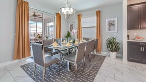 Un coin repas avec une table en verre, des chaises grises et un tapis à motifs. On y trouve également un ventilateur de plafond, un lustre et une plante en pot.
