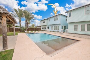 A swimming pool with lounge chairs, a thatched-roof structure, and light blue houses. - Ruby Tuesday Cottage! Community Pool! Downtown PA! (Port Aransas)