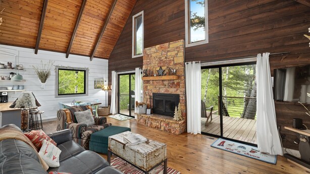 A cozy cabin interior with a stone fireplace, wooden walls, a dining area, and a balcony with a view of the forest.