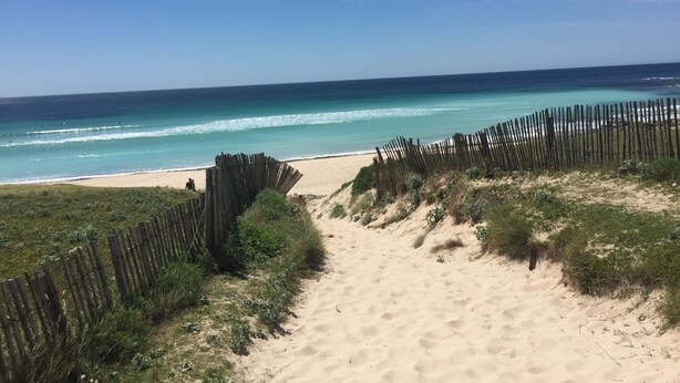 Ein sandiger Strandweg führt zum Meer, gesäumt von einem Holzzaun.