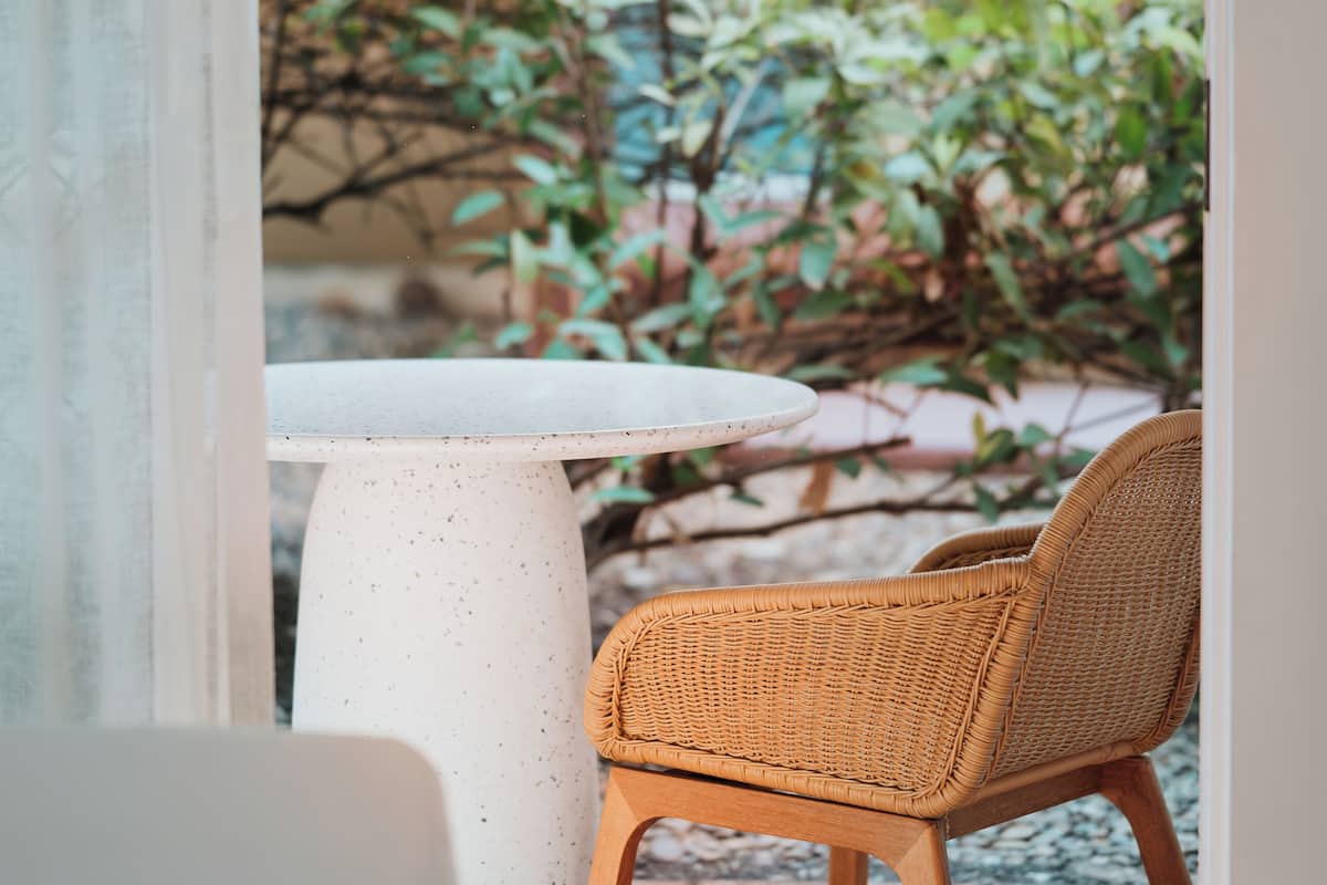 Ein Garten mit runder Tisch und Korbstühlen, einer Betonstütze und Blick auf ein Beet mit verschiedenen Pflanzen.