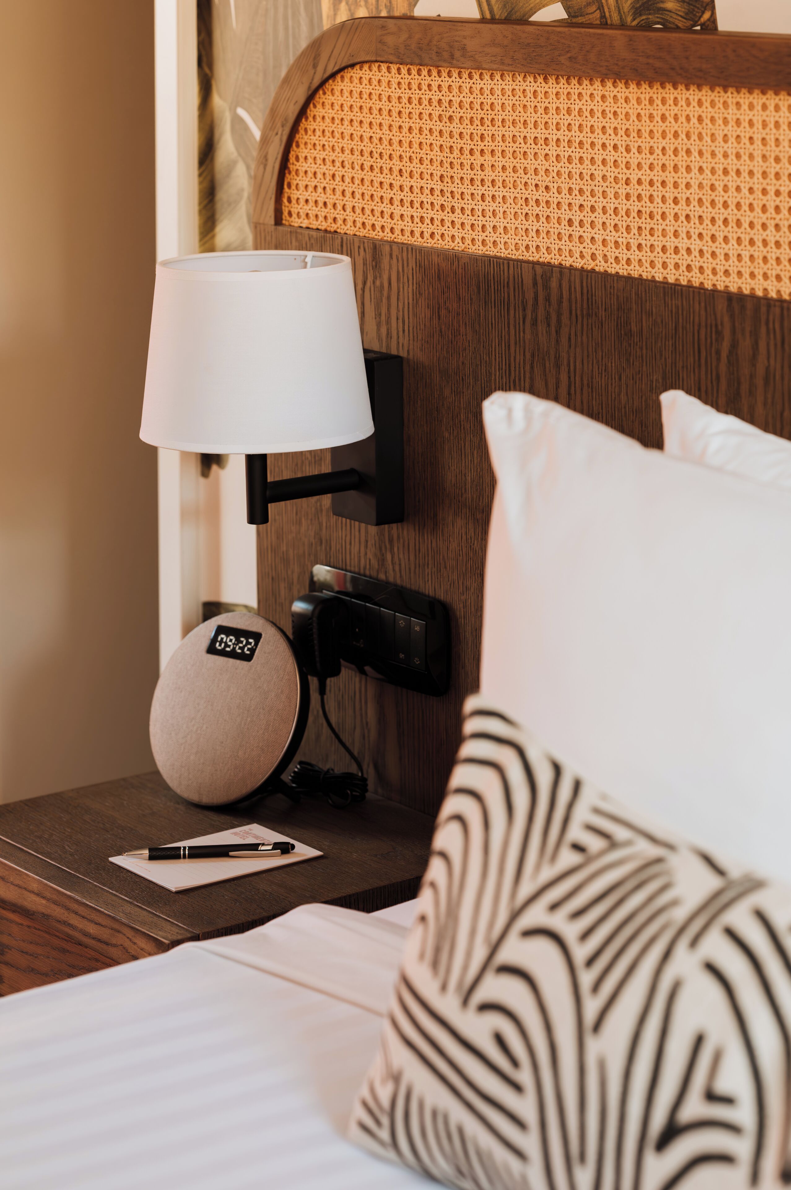 A neatly made hotel room with a bed, a bedside table with a lamp, a clock, and a pillow.