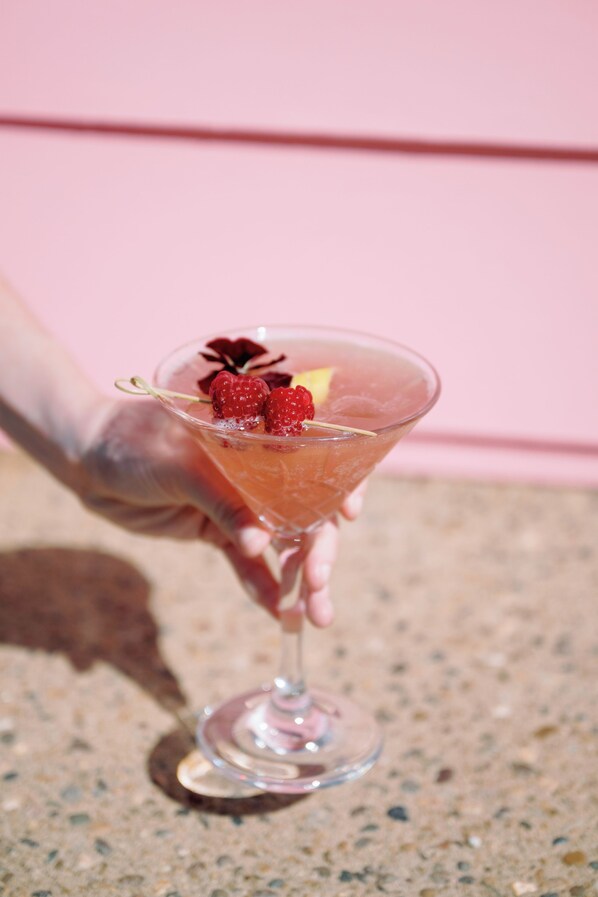 A hand holding a martini glass with a cocktail, garnished with raspberries and a cherry, against a pink background.
