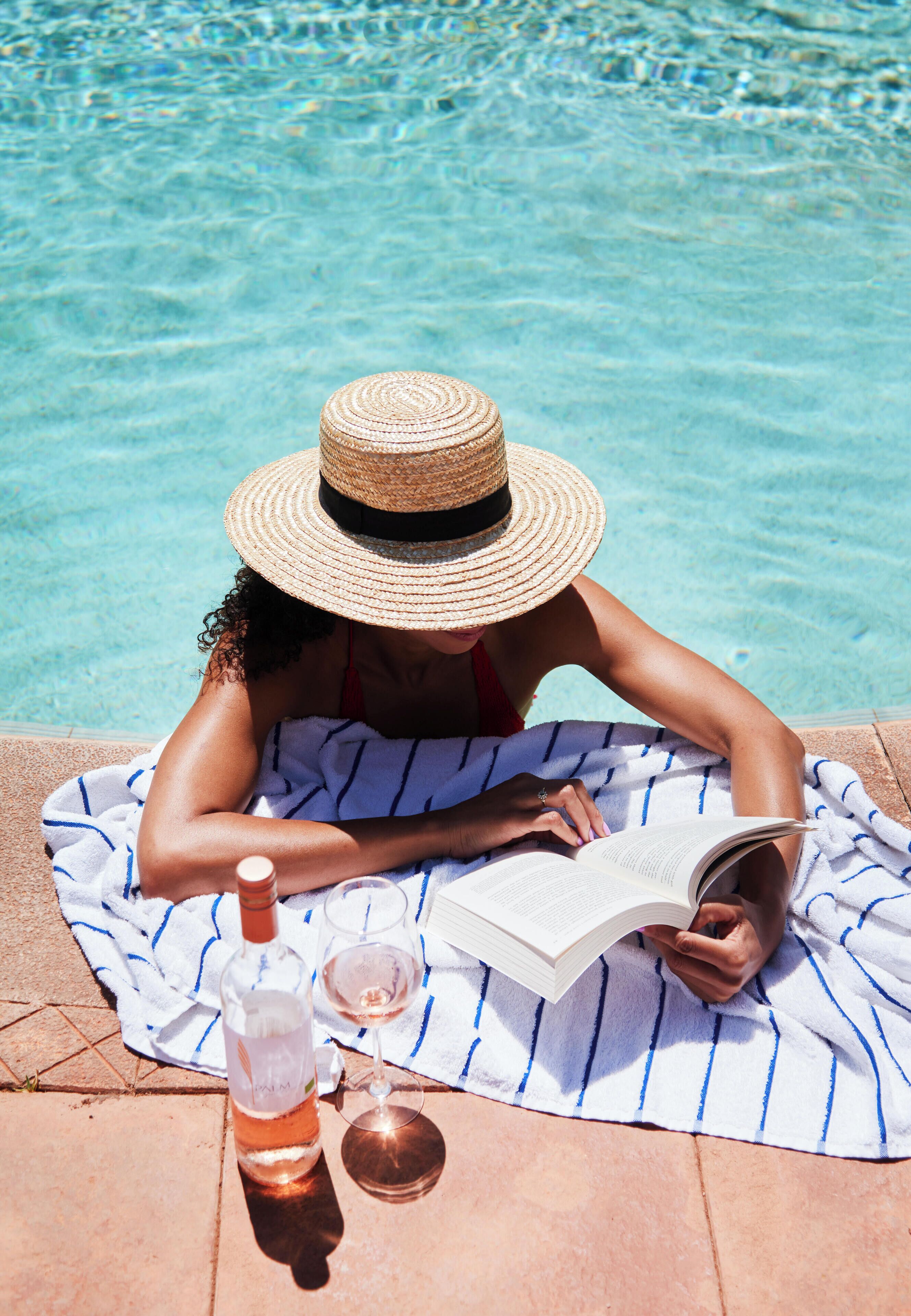 A person reading a book by a poolside with a striped towel and a bottle of wine.