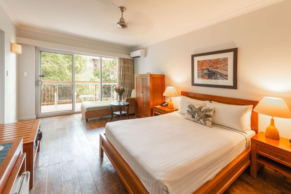 Ein Hotelzimmer mit Bett, Nachttischen, Schreibtisch, Stuhl, Kleiderschrank und einem Balkon mit Blick auf Grünflächen.