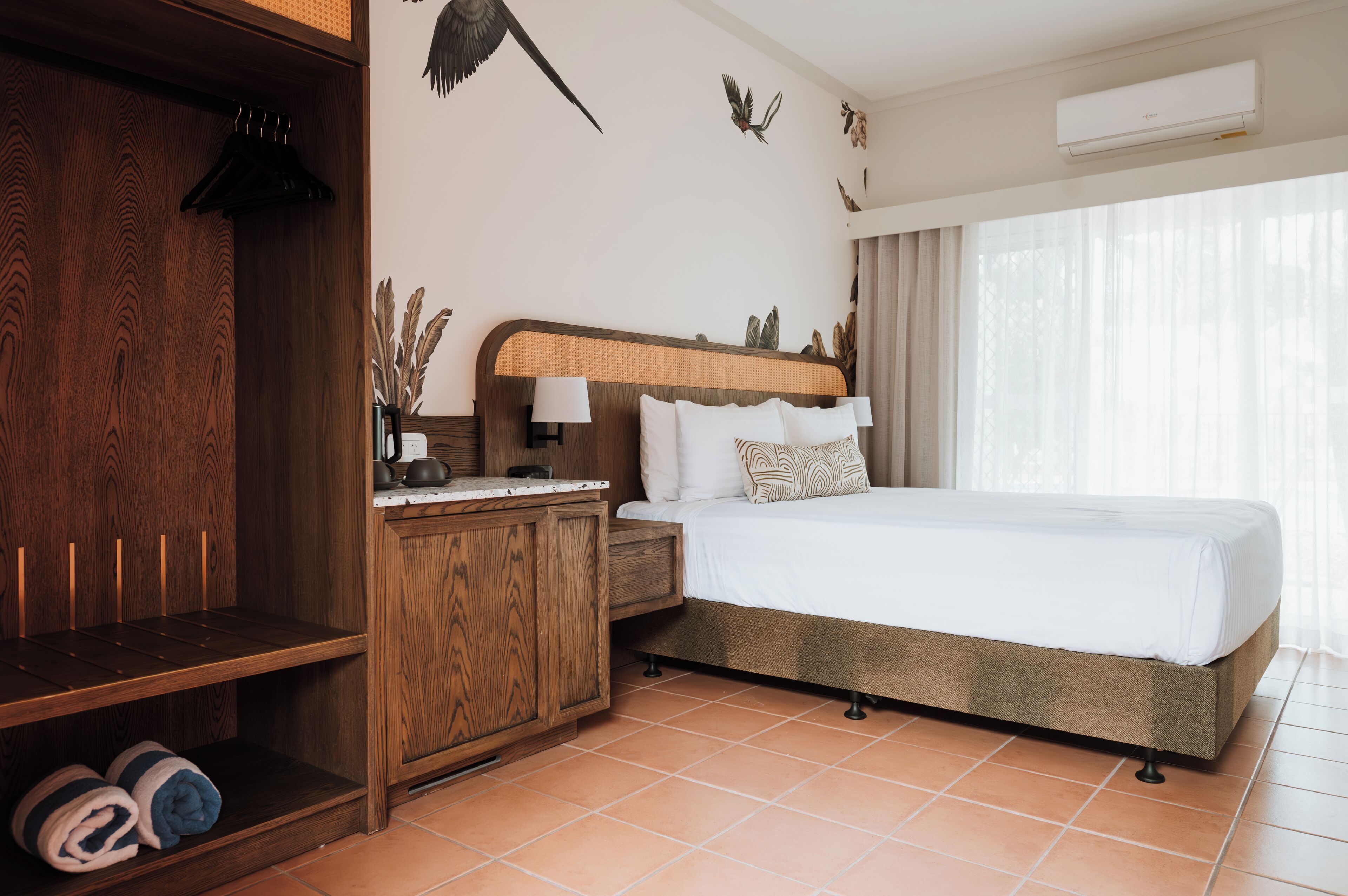 Habitación de hotel con armario de madera, cama con ropa de cama blanca, mesita de noche con lámpara y aire acondicionado instalado en la pared.