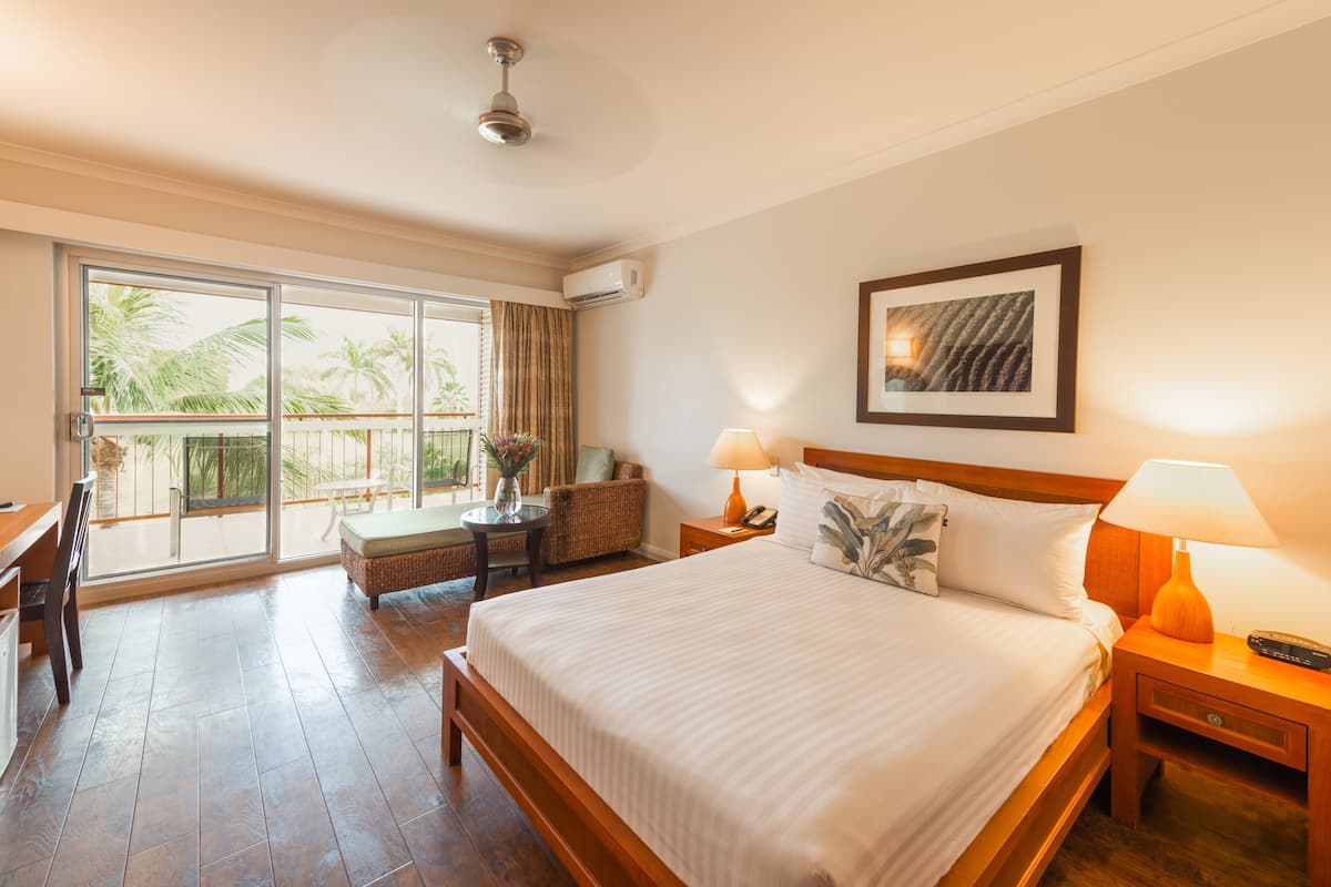 Ein Hotelzimmer mit einem großen Bett, zwei Nachttischen, einem Sessel, einem kleinen Tisch mit einer Vase und einem Balkon mit Blick auf Palmen.