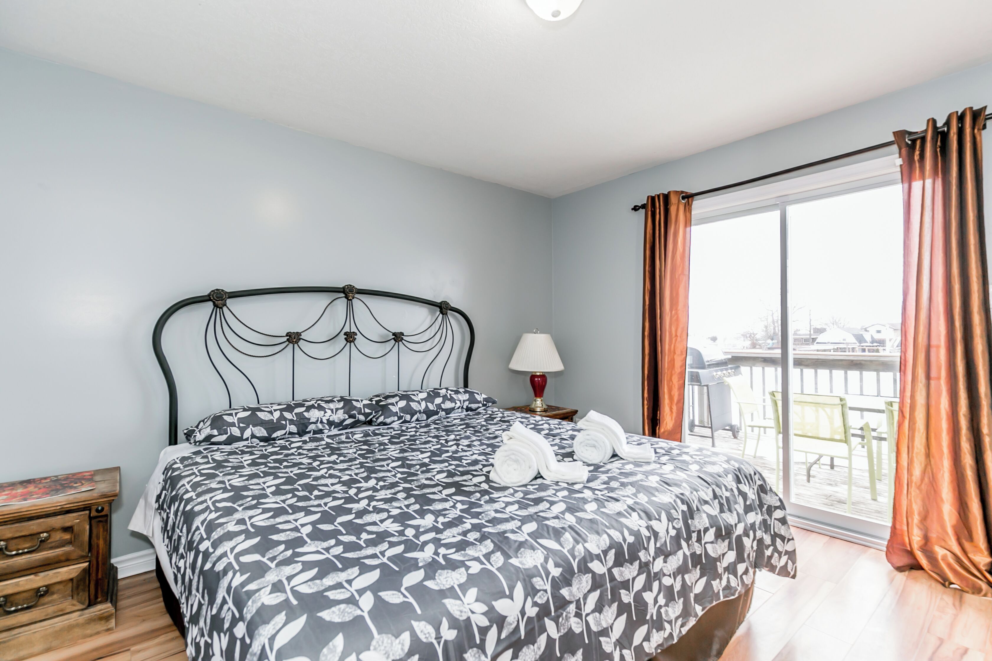 A bedroom with a bed, a nightstand, a lamp, and a view of the outdoors.