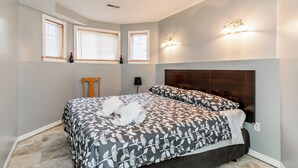 A bedroom with a large bed, two windows with blinds, a nightstand, and a wall-mounted light fixture.