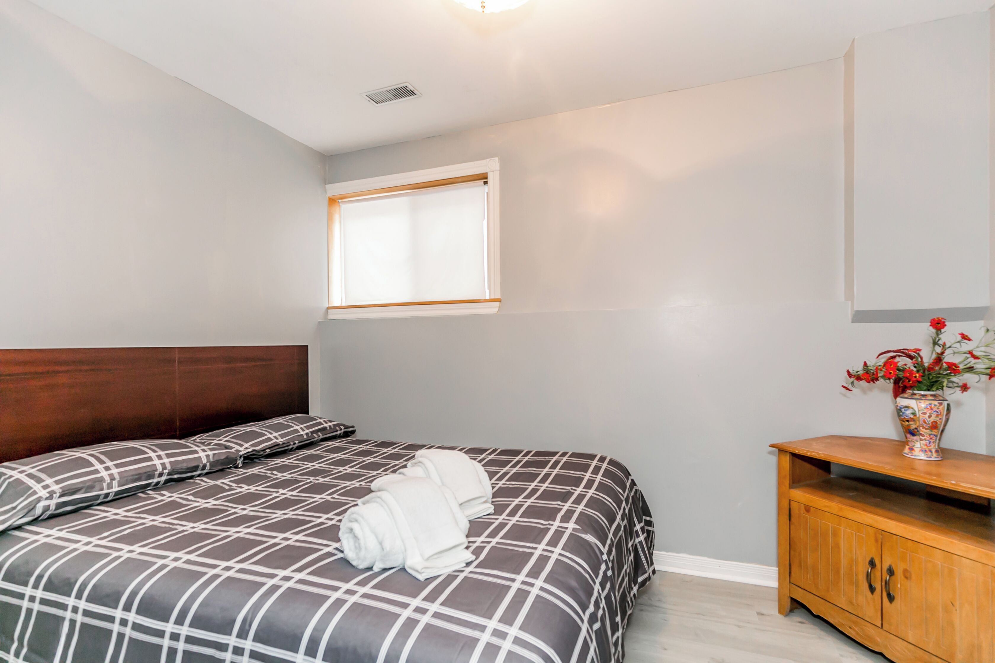 A bedroom with a bed, a wooden headboard, a window, a nightstand, and a vase with flowers.