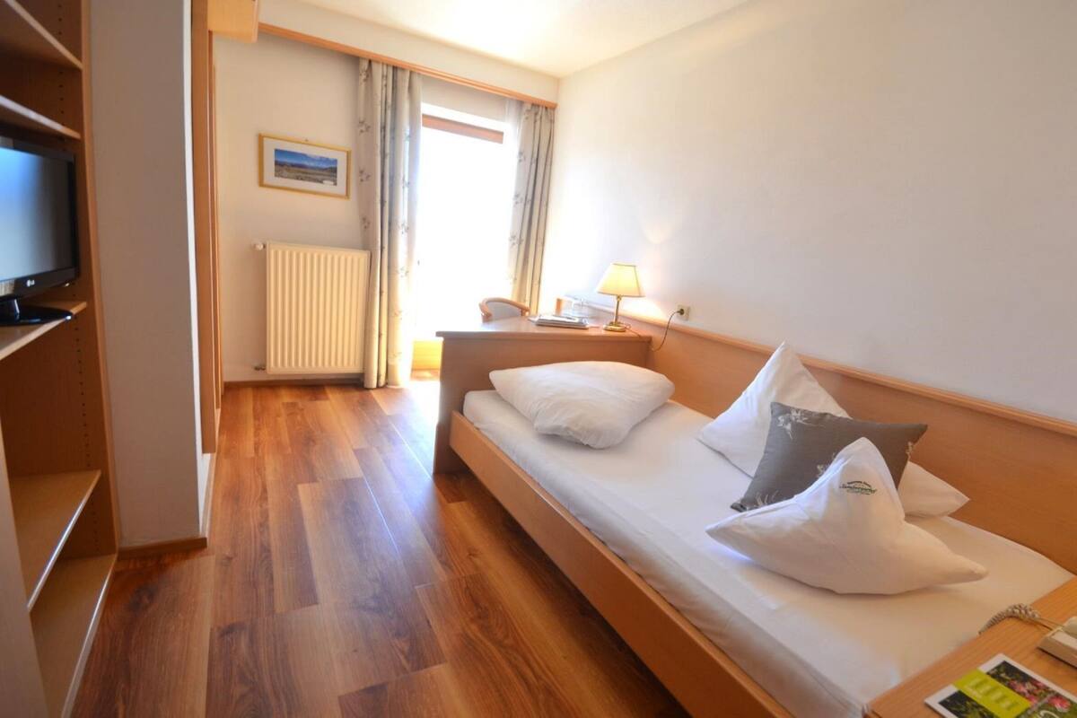 Habitación de hotel con piso de madera, cama con almohadas blancas y grises, escritorio con lámpara, teléfono y vista al exterior a través de una ventana.