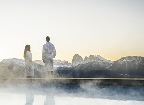 Deux personnes vêtues de robes blanches, debout sur une surface réfléchissante, avec un paysage montagneux en arrière-plan.