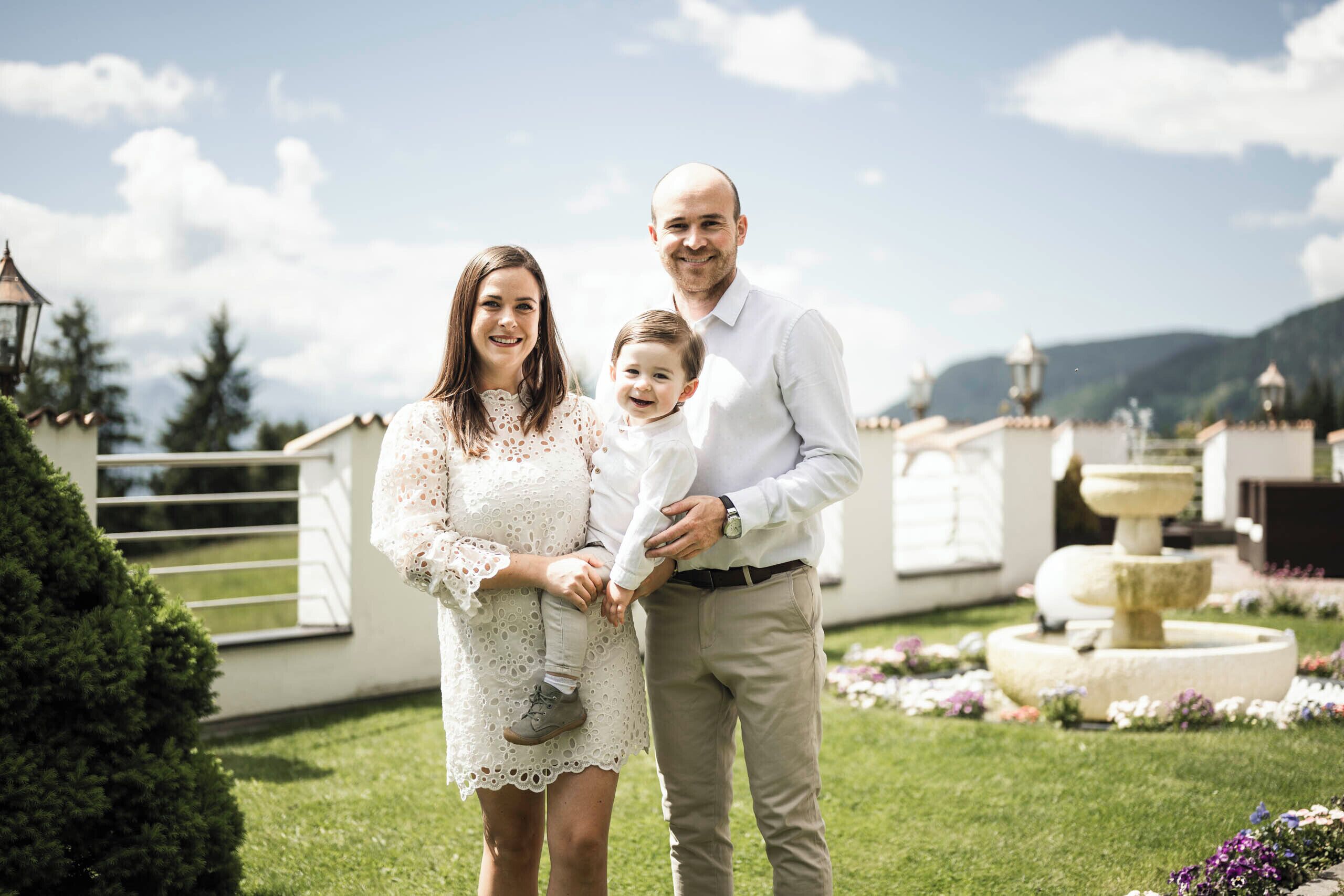 Una familia está de pie en un jardín con una fuente y un edificio al fondo.