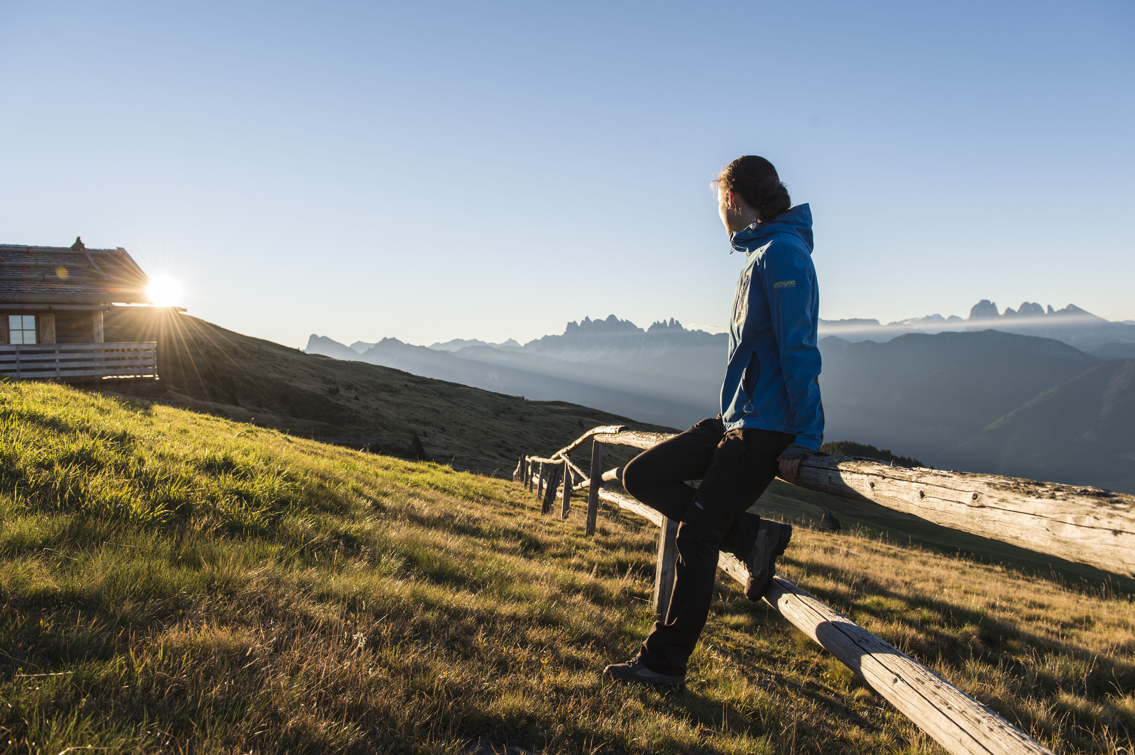 A person sitting on a wooden bench overlooking a mountainous landscape with a cabin in the background.