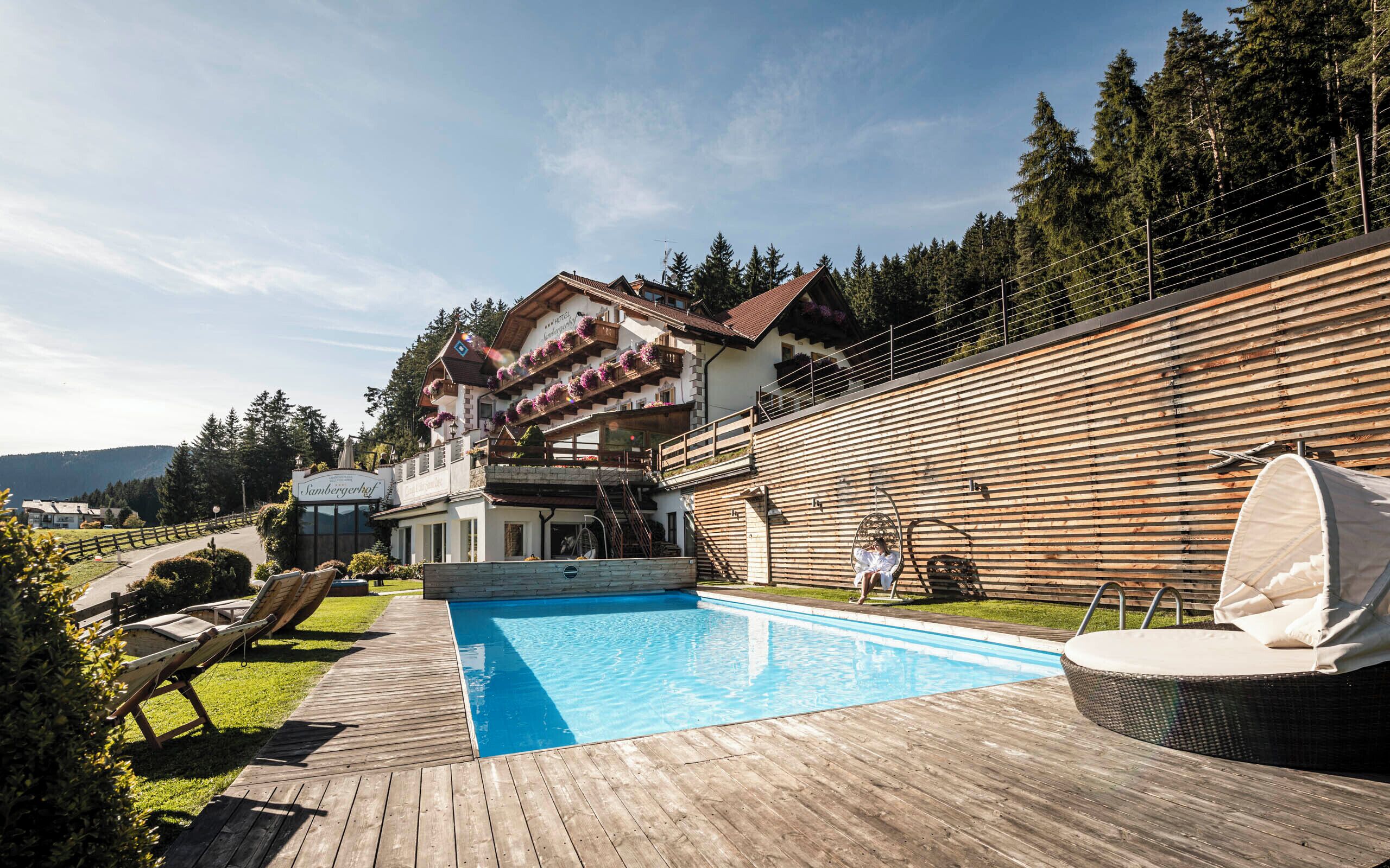 Un hotel con piscina, terraza de madera y montañas al fondo.