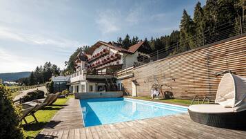 Un hotel con piscina, terraza de madera y montañas al fondo.