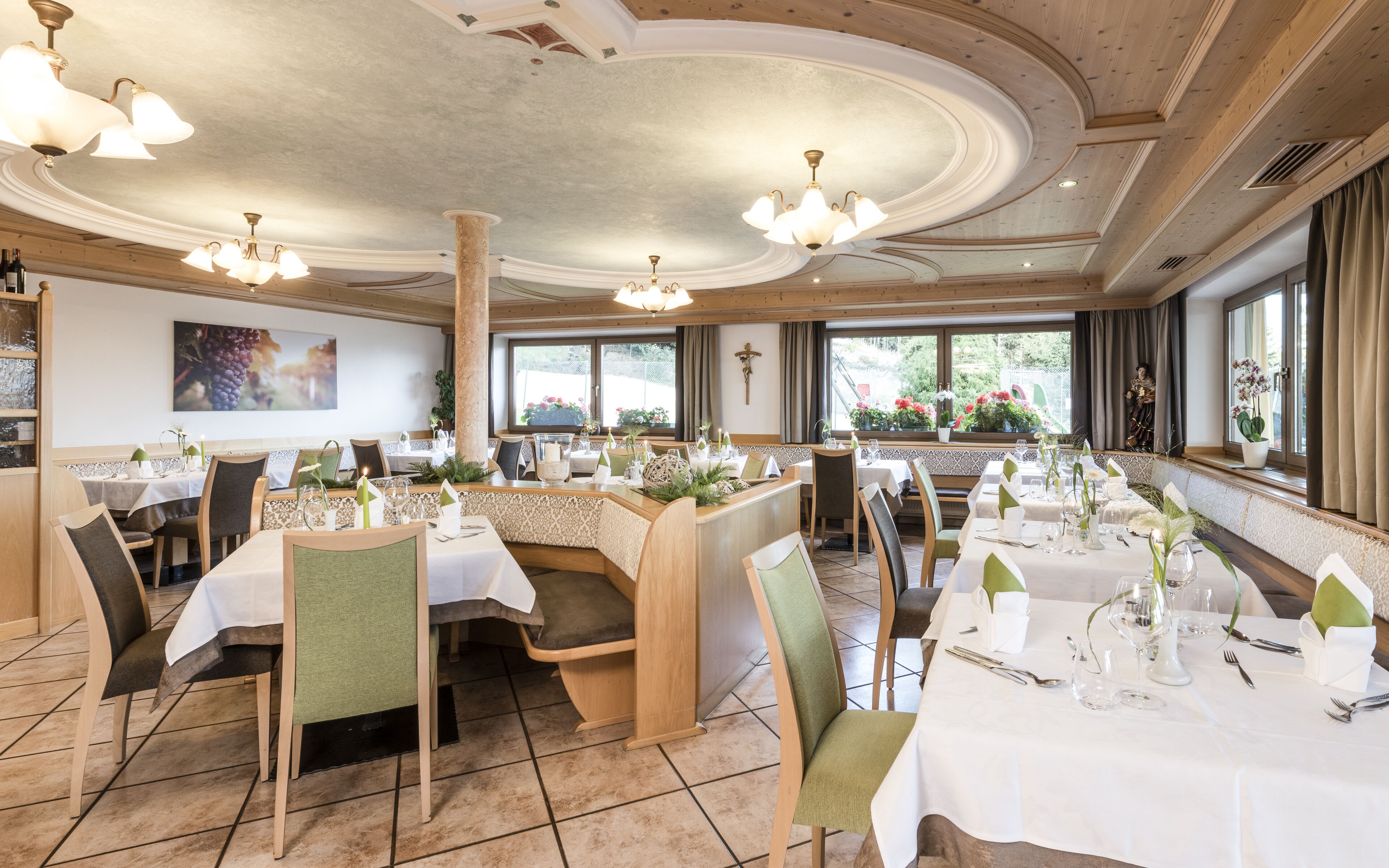 A restaurant with multiple tables set for dining, featuring a high ceiling with chandeliers, large windows with views of greenery, and a cross on the wall.