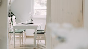 A white dining table with two chairs, a potted plant, and a window with a view of trees.