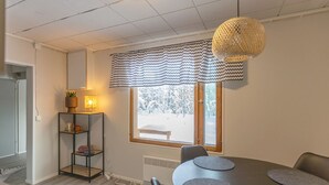 Un comedor moderno con una mesa redonda, sillas grises acolchadas, un tapete blanco y negro estampado, una estantería con objetos decorativos, y una ventana con vista a un paisaje nevado.