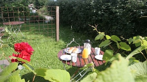 Mesa de jardín con alimentos para el desayuno, rodeada de follaje y una valla metálica.