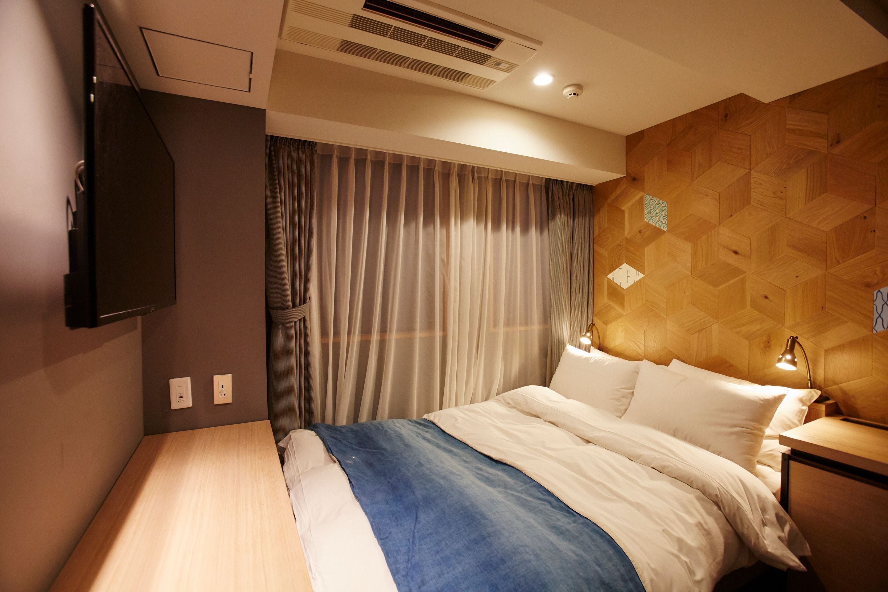 Una camera d'albergo con un letto, una televisione a parete, una testiera in legno e un comodino con una lampada.