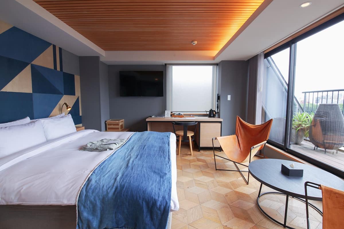 Una habitación de hotel moderna con una cama grande, un escritorio, una silla y vistas de la ciudad a través de amplias ventanas.