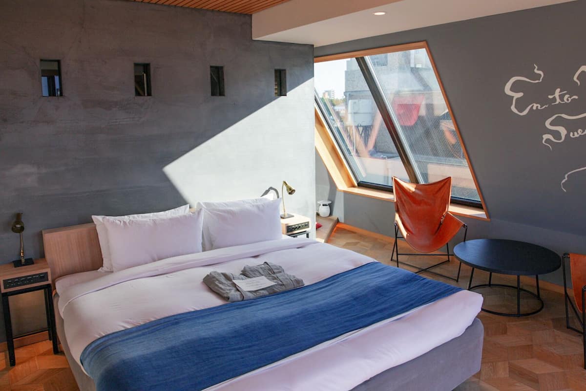 Una habitación de hotel moderna con una cama grande, un tragaluz, una silla roja y una mesita pequeña.