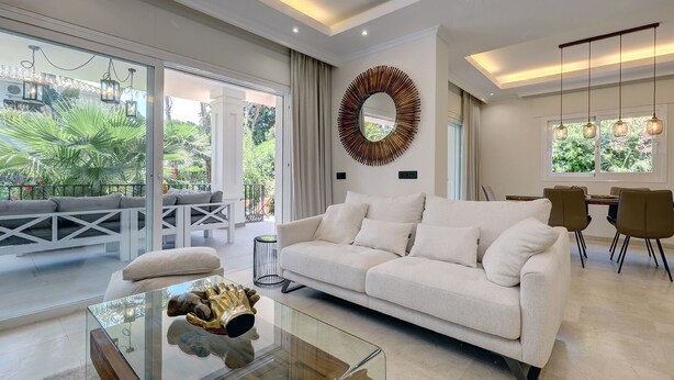 A modern living room with a white sofa, glass coffee table, and a dining area with a table and chairs. - Beautiful 4 Bed Villa ⭐ Heated Pool ⭐ 650m Beach & Dunas de Artola (Marbella)