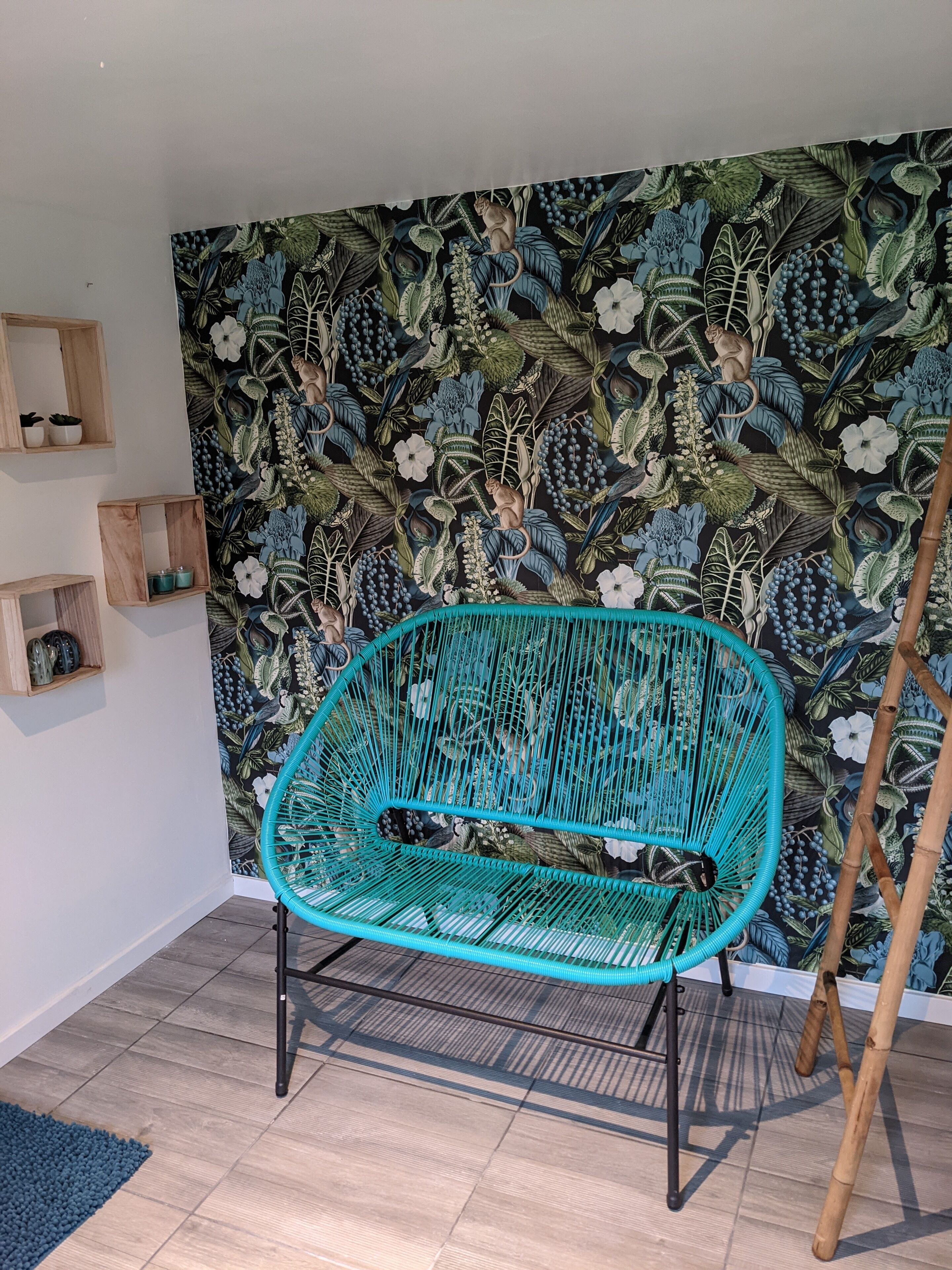 A teal woven chair in a room with a floral wallpaper and wooden shelves.