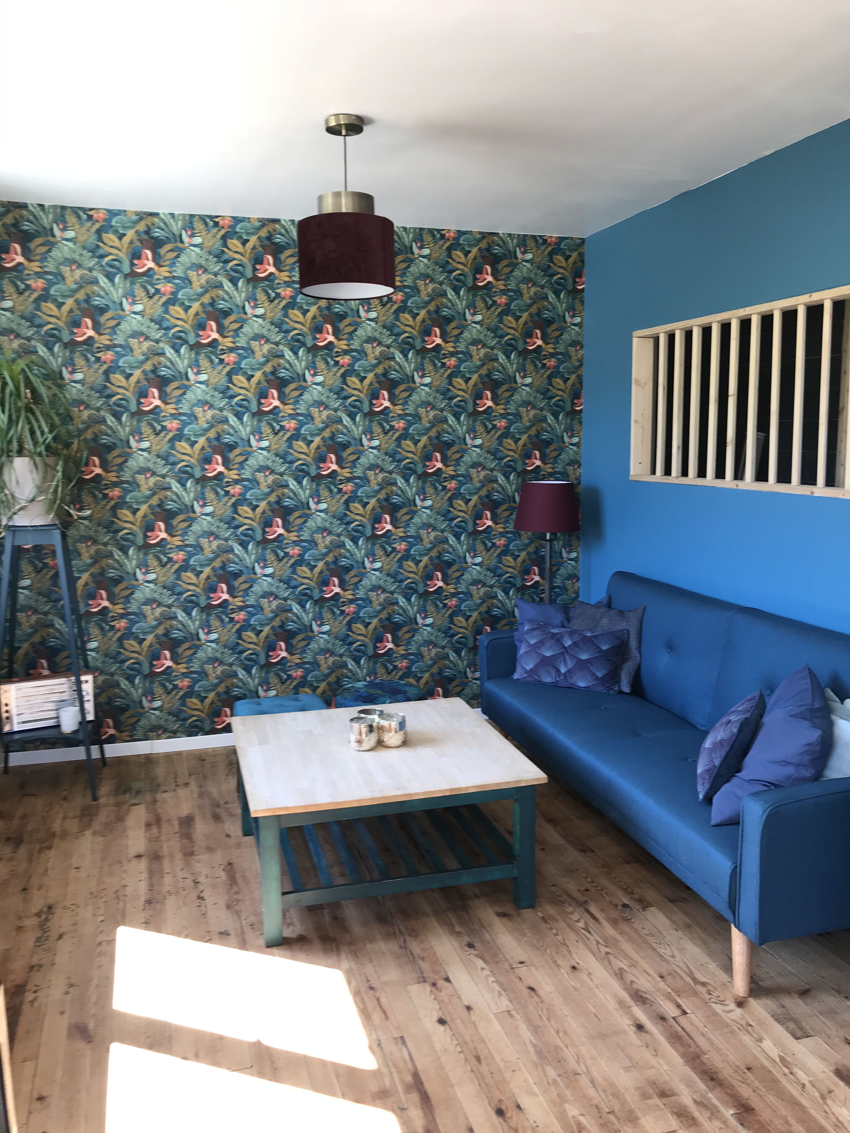 Une pièce avec un canapé bleu, une table basse en bois, un papier peint à motifs, un luminaire suspendu et une fenêtre avec des barreaux.