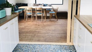 A modern kitchen with a dining area, wooden flooring, and a view of greenery through a large window.