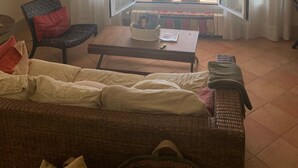 Ein Zimmer mit Blick auf den Strand, ein Korbsessel, ein Tisch mit Zeitschriften und ein Sessel mit rotem Kissen.