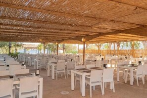 An outdoor dining area with white tables and chairs, a thatched roof, and a view of the sea.