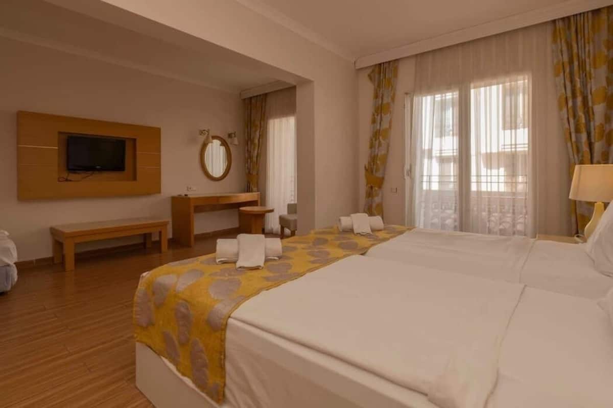 A hotel room with two beds, a TV mounted on the wall, a mirror, a bench, and a window with curtains.