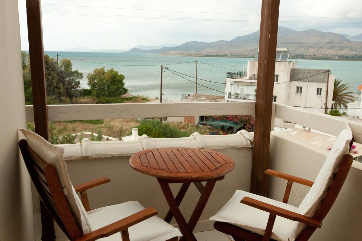 A balcony with two wooden chairs and a small round table, overlooking a body of water and distant mountains.