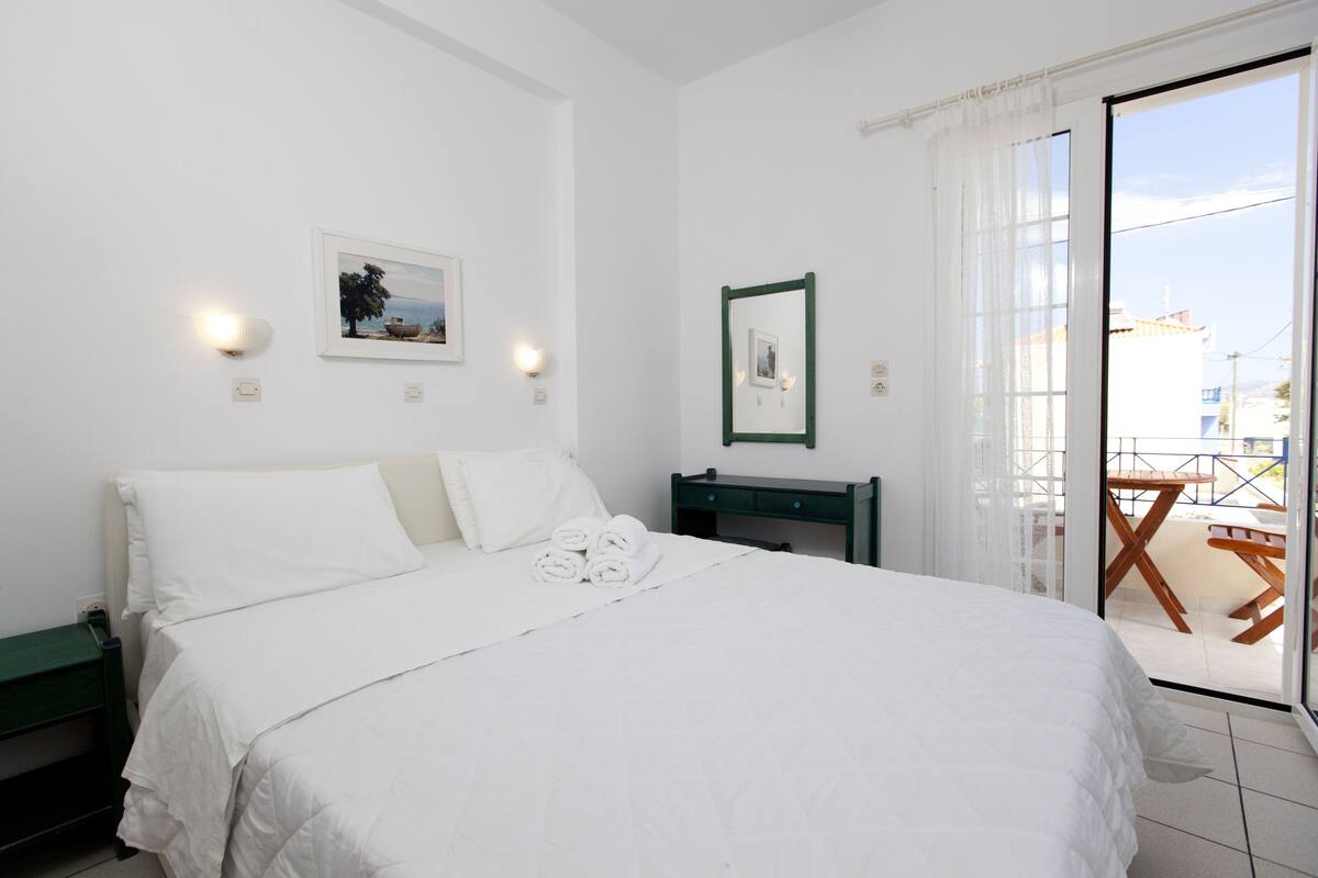 A hotel room with a bed, a nightstand, a small table, and a view of the outside balcony.